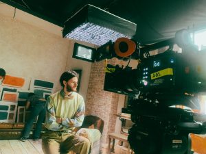 Behind the scenes image from The Phoney lead actor Sam Hume on set, sitting on a stool in front of an Arri Alexa 35 camera and underneath a honeycomb light