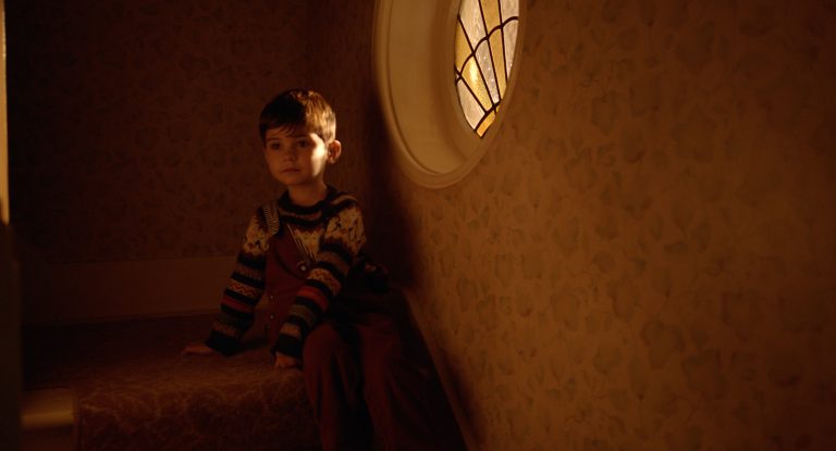 A still image from short film Phonetime depicting a young boy sitting on a staircase. A window to the right illuminates the room with a soft orange glow.