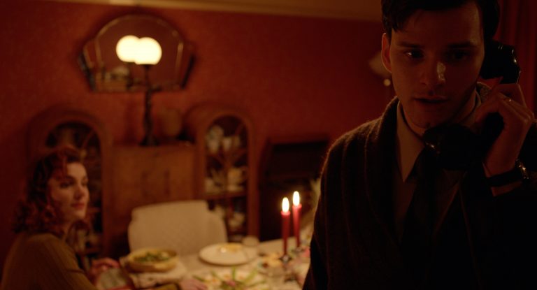 A still image from the short film Phonetime depicting the character Peter holding a rotary dial phone to his ear. His wife sits at a dining table behind him, looking on